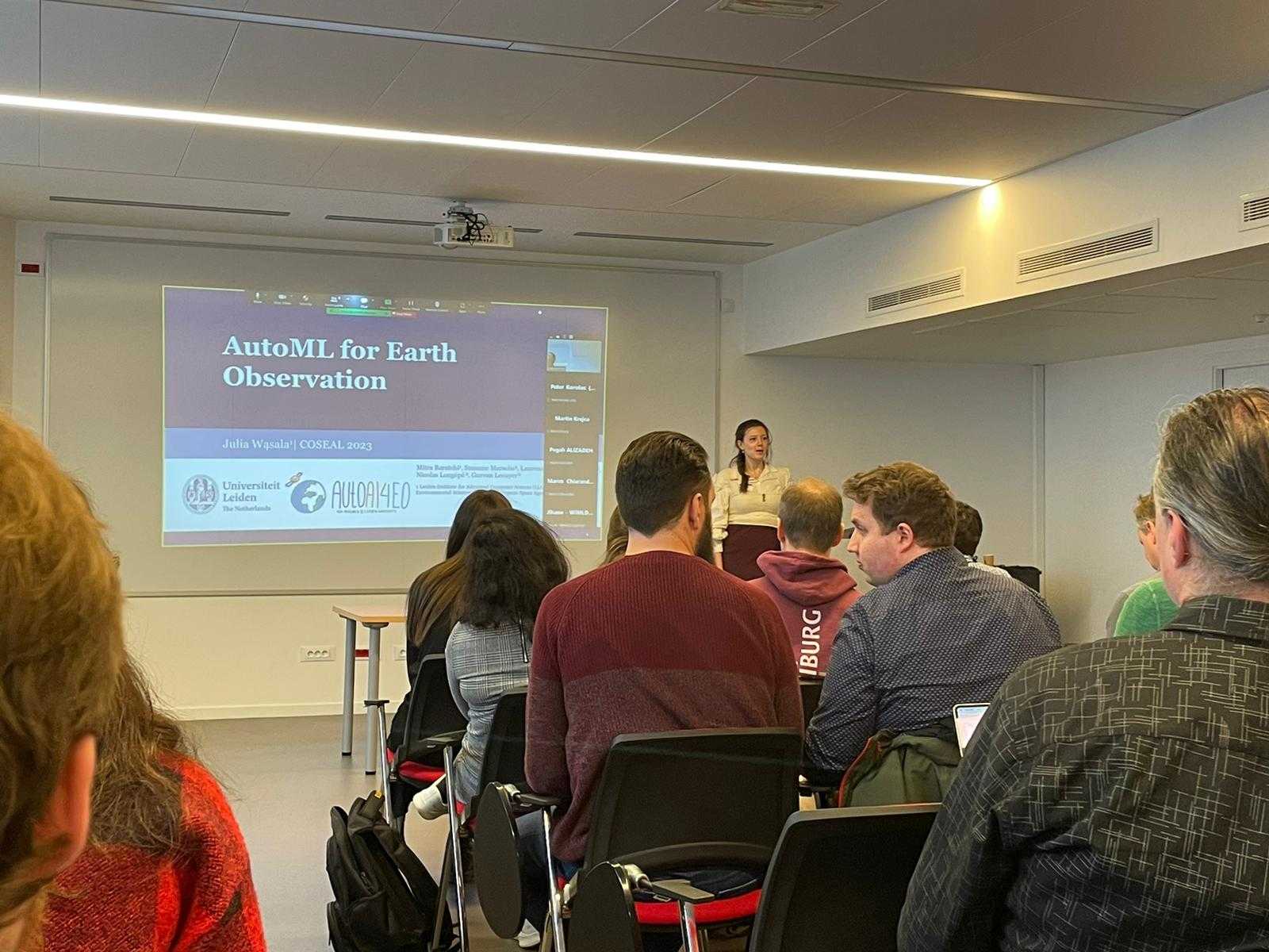 Me presenting my research on AutoML4EO at COSEAL 2023 in Paris. The title slide is projected on the wall. The image is taken from the middle or back from the audience, so you can see a few rows of people. I'm standing at the front of the room, midsentence.